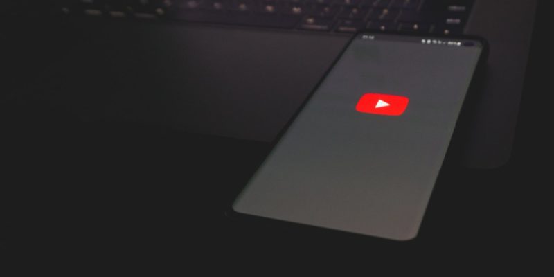 Red youtube logo on a black beackground on phone with a laptop in the background